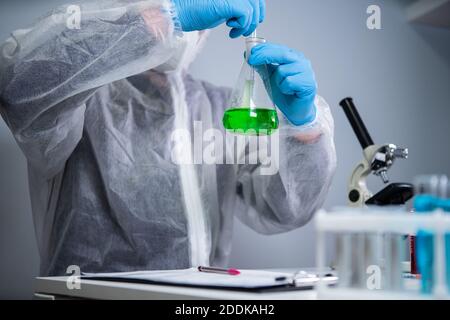 Tecnico di abbigliamento protettivo agitare a mano e agitare liquido verde in matraccio di Erlenmeyer in laboratorio chimico. Studi di tecnici di laboratorio Foto Stock