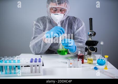 Tecnico di abbigliamento protettivo agitare a mano e agitare liquido verde in matraccio di Erlenmeyer in laboratorio chimico. Studi di tecnici di laboratorio Foto Stock