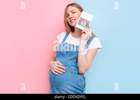 Foto di una giovane donna in stato di gravidanza sorridente e felice che tiene una scansione ecografica mentre si trova su uno sfondo rosa e blu isolato Foto Stock