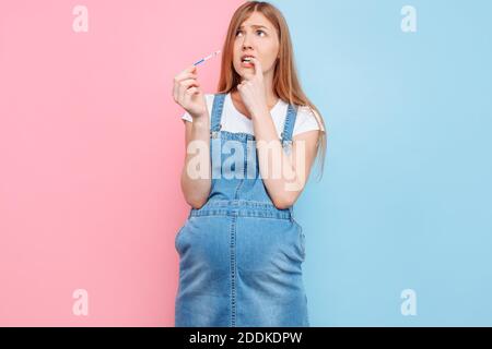 Sì, sono incinta UNA bella donna incinta scioccata mostra un risultato positivo di due strisce su un test, in piedi su una schiena rosa e blu isolata Foto Stock