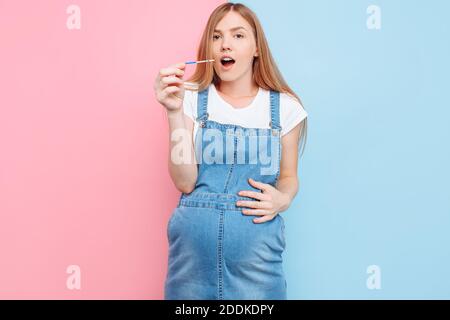 Sì, sono incinta UNA bella donna incinta scioccata mostra un risultato positivo di due strisce su un test, in piedi su una schiena rosa e blu isolata Foto Stock