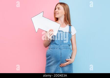 Felice bella giovane donna incinta che tiene un layout di carta vuoto sotto forma di una freccia che punta ad una copia dello spazio sullo sfondo, in piedi Foto Stock