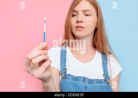 Sì, sono incinta UNA bella donna in gravidanza felice mostra un risultato positivo di due strisce sul test, in piedi su una parte posteriore isolata rosa e blu Foto Stock