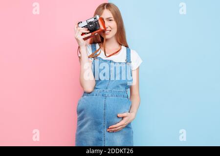 Bella giovane donna incinta in denim vestito scattando foto sopra telecamera retrò isolata su sfondo rosa e blu Foto Stock