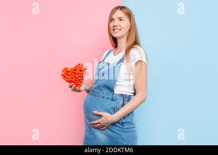 Felice bella donna incinta che tiene un cuore rosso vicino al ventre, in piedi su uno sfondo rosa e blu Foto Stock