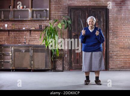 Donna anziana allegra che tiene coltelli da cucina all'interno. Concetto di sicurezza e autodifesa. Potrebbe essere usato in modo umoristico. Foto Stock