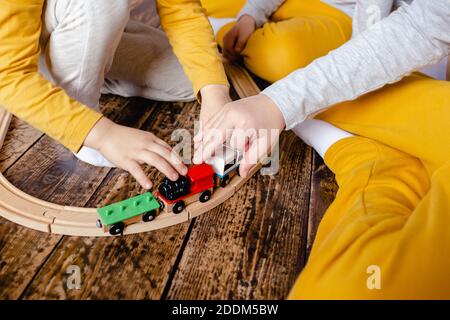 I bambini piccoli costruiscono la ferrovia e giocano con il treno di legno seduto sul pavimento nel soggiorno. Bambini piccoli che giocano con un'auto giocattolo. I bambini giocano. Foto Stock