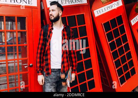 Uomo elegante con barba, hipster vestito con una T-shirt bianca e una camicia a quadri, uomo che tiene uno skateboard nelle mani e che posa davanti a una t rossa Foto Stock