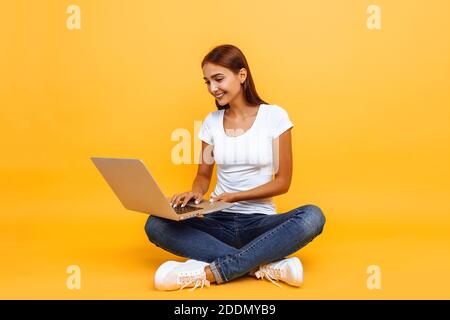 Ritratto di giovane donna sorridente seduto a gambe incrociate, utilizzando un computer portatile isolato su sfondo giallo Foto Stock