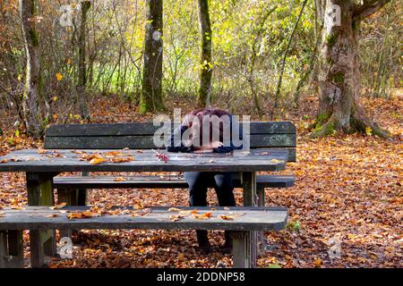 Donna con i capelli scuri seduta ad un tavolo da picnic con la testa giù sulle sue mani. Foglie d'autunno che si posano ovunque. Foto Stock