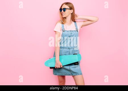 Felice elegante giovane ragazza in occhiali da sole, con uno skateboard in posa su uno sfondo rosa isolato Foto Stock