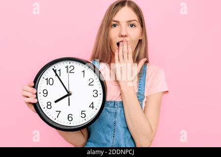 Scioccato, una giovane donna su uno sfondo rosa isolato che tiene un orologio da parete. Foto Stock