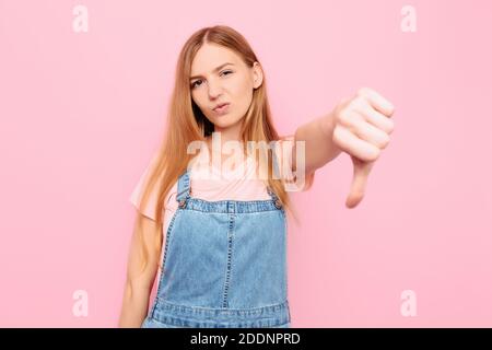 Dissimile, Ritratto di una giovane ragazza scontenta che fa un gesto di pollice giù esprimendo malcontento, disapprovazione, critica, su uno sfondo rosa Foto Stock