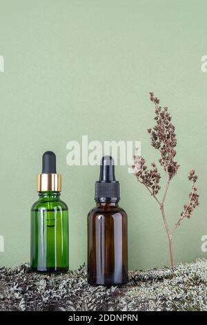 Due bottiglie di vetro marrone verde con siero, olio essenziale o altro prodotto cosmetico e piante di fiori secchi su corteccia di alberi ricoperte di muschio contro la b verde Foto Stock