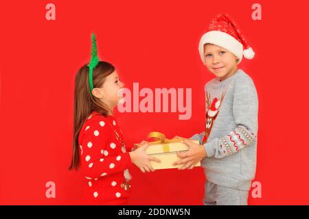 Due bambini in pullover hanno in mano un regalo di Capodanno su sfondo rosso. Un ragazzo e una ragazza danno un regalo di Natale a vicenda Foto Stock