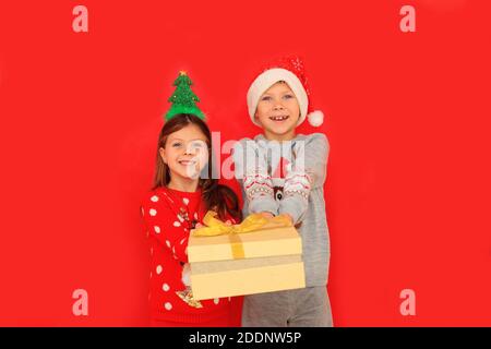 Due bambini in pullover hanno in mano un regalo di Capodanno su sfondo rosso. Un ragazzo e una ragazza danno un regalo di Natale a vicenda Foto Stock