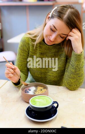 Frullato ciotola di cibo crudo e latte matcha tazza di tè verde sul tavolo e una donna pronta a colazione. Foto verticale Foto Stock