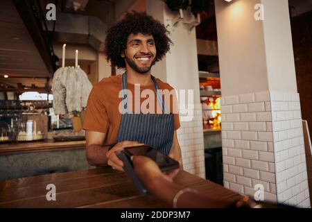 Barista giovane uomo che indossa abiti casual con grembiule senza contanti il terminale deve pagare l'ordine al banco per il cliente tramite smartphone Foto Stock