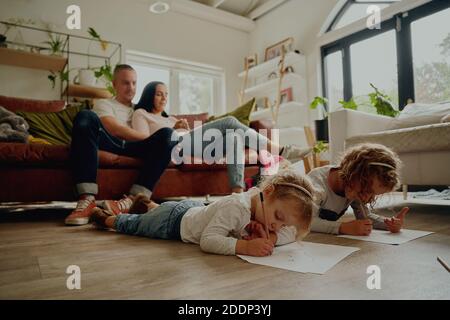 I genitori seduti sul divano con due belle figlie carine mentite sul pavimento che crea opere d'arte su carta a casa Foto Stock