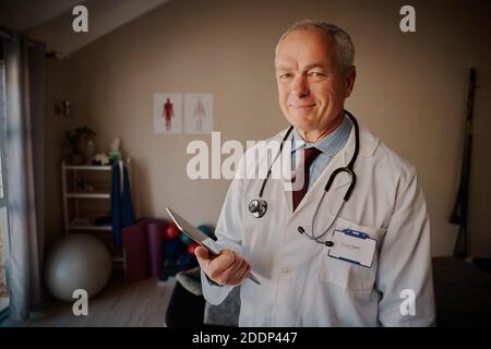 Ritratto di un medico senior sorridente di successo che tiene in mano un tablet digitale alla telecamera Foto Stock