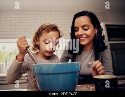 Madre felice insegnando la bambina che si cuoce in cucina e si mescola impasti nel recipiente Foto Stock