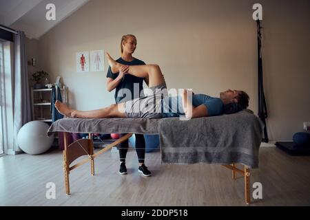 Giovane fisioterapista femminile che diagnostica le lesioni al ginocchio dei pazienti di sesso maschile durante il massaggio letto Foto Stock
