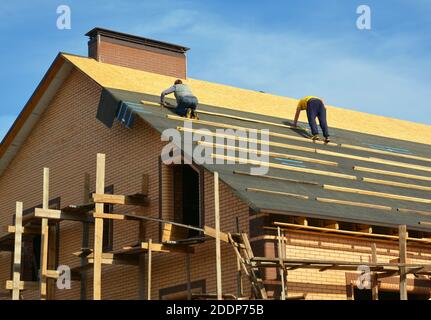 I fornitori di coperture stanno installando underlayment, membrana impermeabile su un ponte di tetto, rivestimento di compensato di una grande casa di mattoni sotto costruzione wi Foto Stock