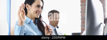 Donna d'affari sorridente si siede allo schermo del computer con microfono e onde consegnare il collega in background in ufficio Foto Stock
