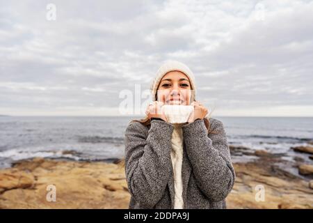 Bella giovane donna caucasica stringe il collo del suo bianco maglione con le mani per il freddo inverno sorridente guardando la macchina fotografica sul mare Foto Stock