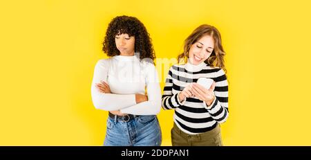 Due giovani donne di razza mista isolato divertente immagine sul giallo Sfondo per la copia spazio - donna caucasica che si diverte con Smartphone mentre il suo nero Foto Stock