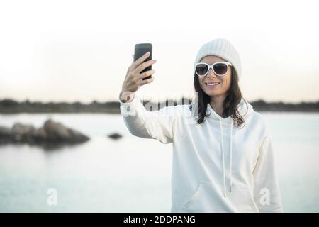 Giovane bella donna con grandi divertenti occhiali da sole bianchi in piedi una riva del lago che fa un ritratto di sé per condividerla vacanza invernale con i suoi amici Foto Stock