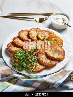 Frittelle di patate con formaggio di cottage con aglio e prezzemolo, servite con salsa di panna acida su un piatto su uno sfondo di pietra di marmo chiaro con posate dorate Foto Stock