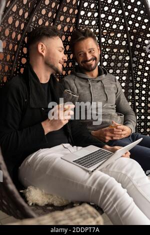 Giovane bella coppia gay ridendo e rilassandosi sull'altalena sul balcone con birra e computer portatile. Foto di alta qualità Foto Stock