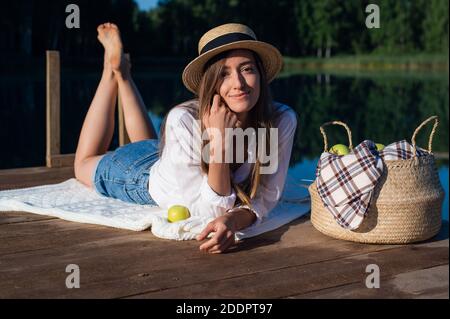 Giovane bella donna su un pic-nic presso il lago, sdraiata su un molo di legno. Indossa una camicia e un paio di pantaloncini in denim. Nelle vicinanze troverete un cesto di frutta e una mela. Foto Stock