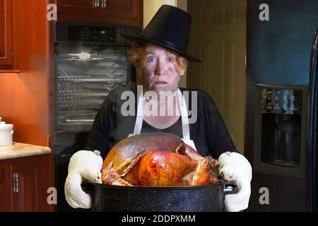 Donna in piedi in cucina indossando pellegrino hat e grembiule con bruciature viso e capelli tenendo una teglia. Foto Stock