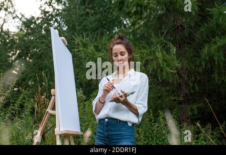 donna artista estate nel parco, sfondo foresta cespugli alberi, disegna foto, vista anteriore, la sua mano palette con vernici, pennello e cavalletto. Jeans vestiti Foto Stock