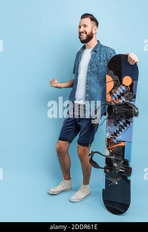 Un uomo felice con la barba , indossando pantaloncini e una camicia , tiene uno snowboard e mostra un gesto di vittoria e di successo, in piedi su un isolato bac blu Foto Stock