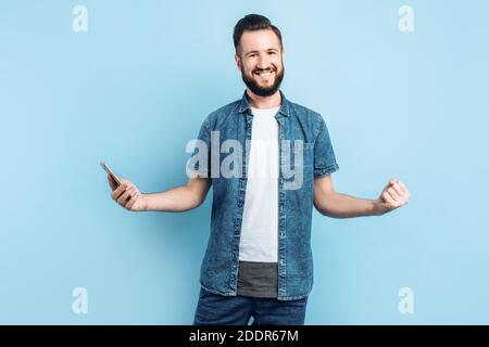 Un uomo sorridente felice con la barba , tenendo uno smartphone in mano e dimostrando un gesto di vittoria e di successo, si erge su uno sfondo blu Foto Stock