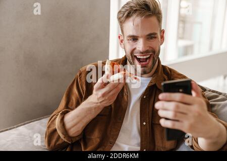 Foto di un bel giovane allegro seduto sul divano al coperto a casa e mangiare la pizza mentre si parla da videocall vicino telefono cellulare Foto Stock