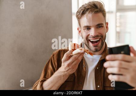 Immagine di sorridente ottimista bel giovane seduto sul divano all'interno a casa e mangiare pizza mentre si parla da videocall tramite telefono cellulare Foto Stock