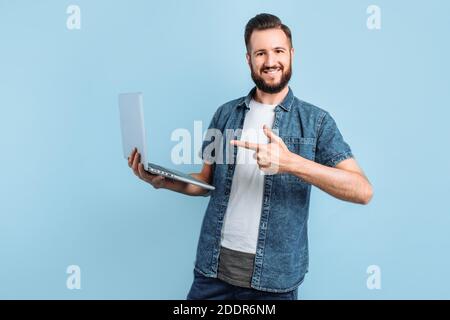 Un uomo felice tiene un laptop e lo punta con il dito, in piedi su uno sfondo blu isolato Foto Stock