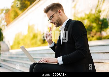Uomo d'affari serio che punta il dito verso l'alto e utilizza il computer portatile mentre si è seduti su scale all'aperto Foto Stock
