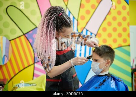 Donna parrucchiere in una maschera faccia fa un taglio di capelli per un ragazzo adolescente in un barbiere per bambini Foto Stock