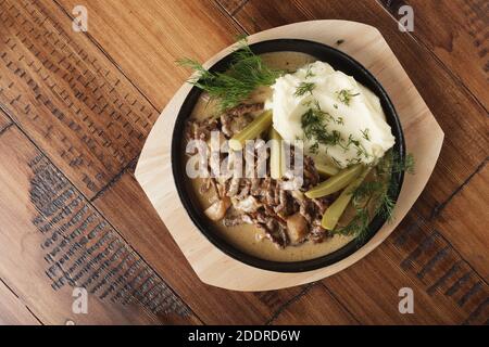 Stroganoff di manzo con patate a fungo e cetrioli sottaceto in una padella. Sfondo di legno Foto Stock