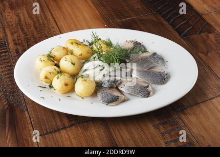 Pesce di aringa con palline di patate giovani su un piatto bianco. Sfondo di legno. Foto Stock