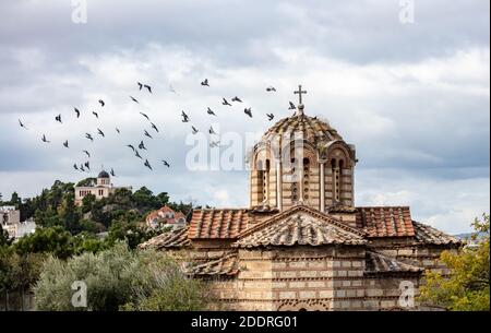 Gregge di piccioni che volano intorno alla chiesa greco-ortodossa nella zona di Thissio, Atene, Grecia. Edificio dell'osservatorio nazionale e sfondo del cielo nuvoloso. Foto Stock