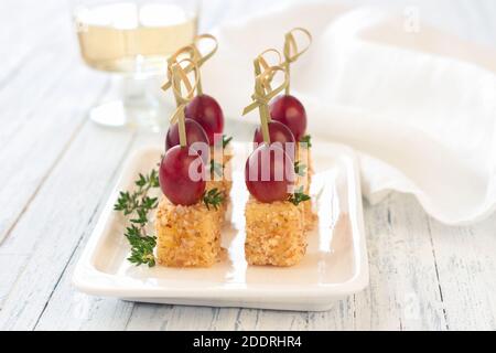 Canape con uva rosa e formaggio in noce impanata, su spiedini di legno. Spuntino festivo su un piatto bianco su sfondo di legno azzurro, selettivo Foto Stock