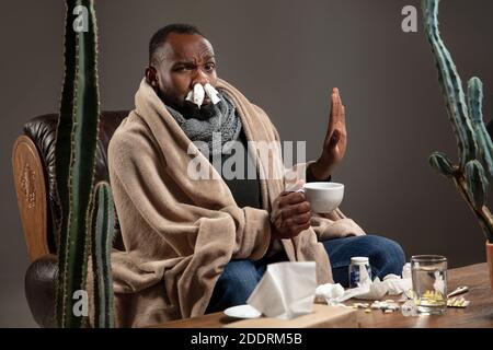 Giovane afroamericano avvolto in una plaid che cerca di proteggere da qualcuno ammalato, sembra disgustato, arrabbiato, triste seduta in poltrona a casa al chiuso. Sanità e medicina, prevenzione dei malati. Foto Stock