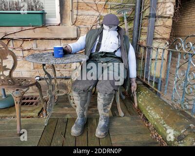 Uomo fittizio di dimensioni standard seduto su sedia con tavolo esterno casa in cappellino piatto pantaloni impermeabili wellingtons con vaso blu di tè e ringhiere di metallo a lato Foto Stock