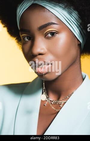 giovane donna afroamericana in abito blu elegante con collana in bocca isolata in giallo Foto Stock